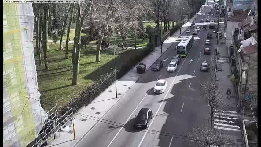 A sunny day shows a city street with several cars and a bus driving on a road that passes a park with leafless trees and a building under construction.