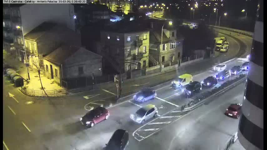 A nighttime, wet street intersection with several cars at a four-way stop, surrounded by buildings.