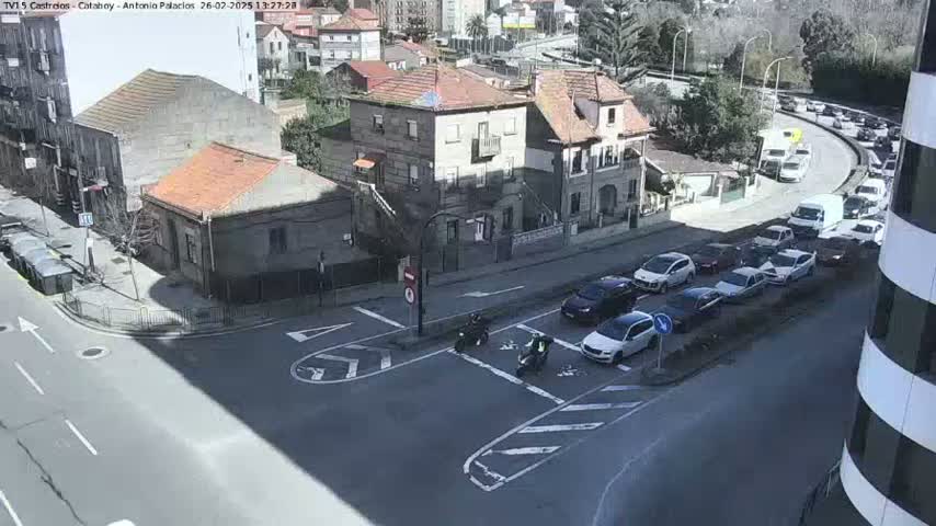 A sunny day reveals a street intersection with several cars stopped in traffic, motorcycles at a crosswalk, and buildings nearby.