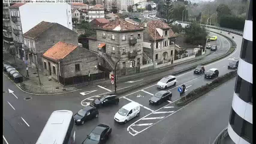 A wet city intersection with several cars and a bus, surrounded by buildings and a curving roadway.