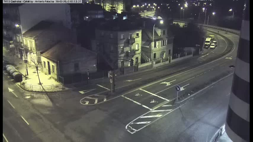 A nighttime, overhead view of a street intersection with several buildings, some cars on a nearby highway, and street markings indicating bike lanes.