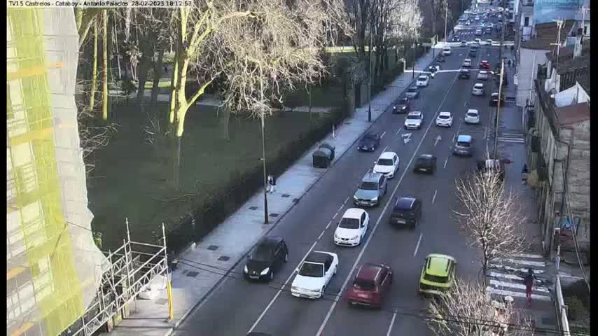 A sunny day shows a multi-lane road with moderate traffic flowing in both directions, flanked by a sidewalk with sparse pedestrians, trees, and buildings.