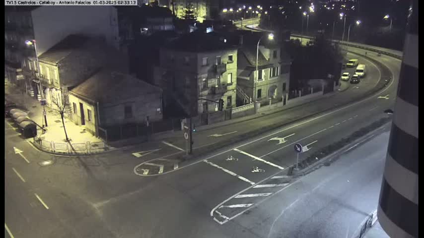 A nighttime, overhead view of a street intersection with several buildings and a few vehicles driving on a curved road.