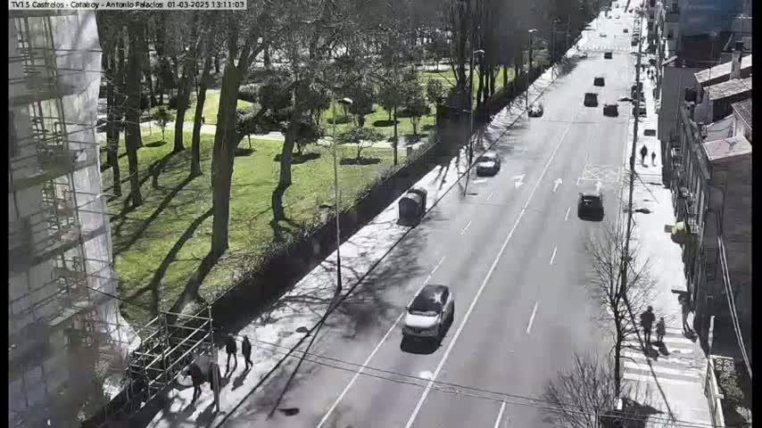 A sunny day shows a city street with several cars driving in both directions, pedestrians walking on sidewalks alongside a park with many trees, and a building under construction.
