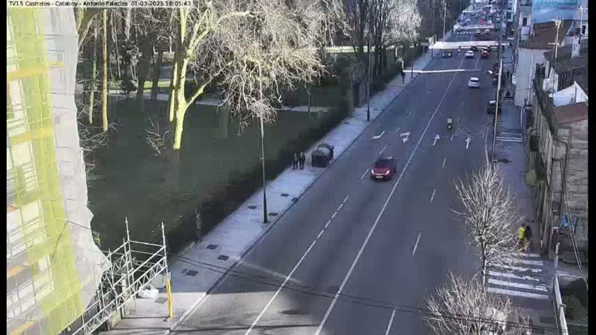 A sunny day reveals a multi-lane road with sparse traffic, flanked by buildings and a park with bare trees.