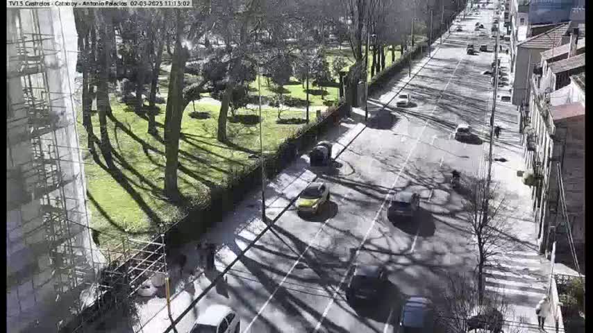 A sunny day shows a city street with cars driving past a park with tall trees casting shadows, next to a building under construction.