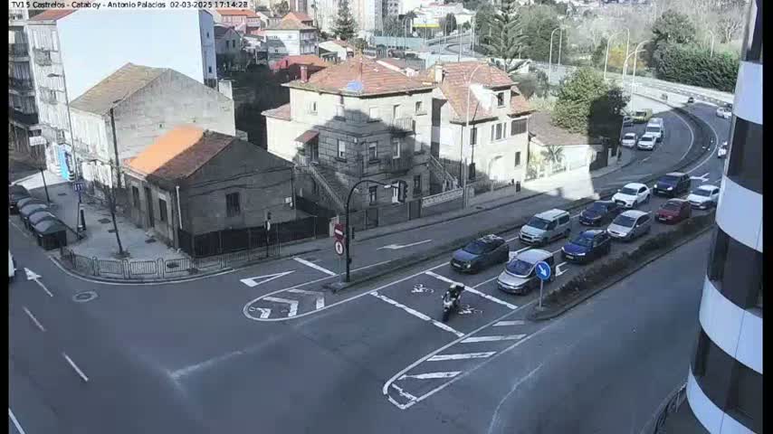 A sunny day shows a city intersection with several cars stopped at a crosswalk, a motorcycle in the middle of the intersection, and multiple buildings in the background.