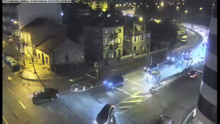 A nighttime street intersection with several cars, some moving and others stopped, is illuminated by streetlights under wet conditions.