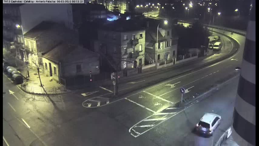 A wet, nighttime street intersection shows a white car at a stop sign near several buildings, with a curved road visible in the background.