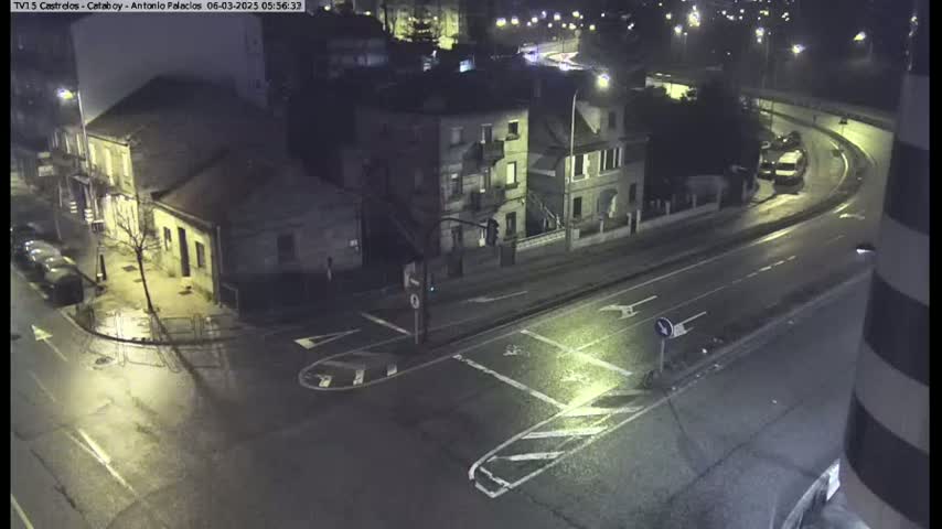 A rain-slicked intersection at night shows several buildings and parked cars, with a curved road visible in the background.