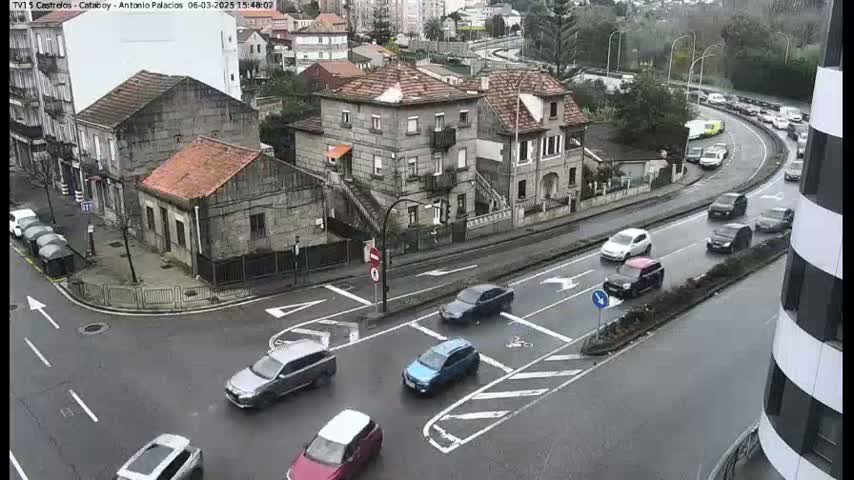 A wet street intersection shows several cars at a crossroads and a nearby road with moderate traffic, all surrounded by stone buildings in a residential area.