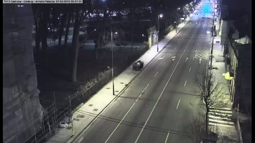 A nighttime, high-angle view shows a mostly empty road lined with trees and buildings, with a single vehicle parked on the side.