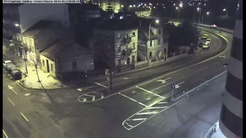 A nighttime, high-angle view shows a street intersection with buildings and cars on a curving road.