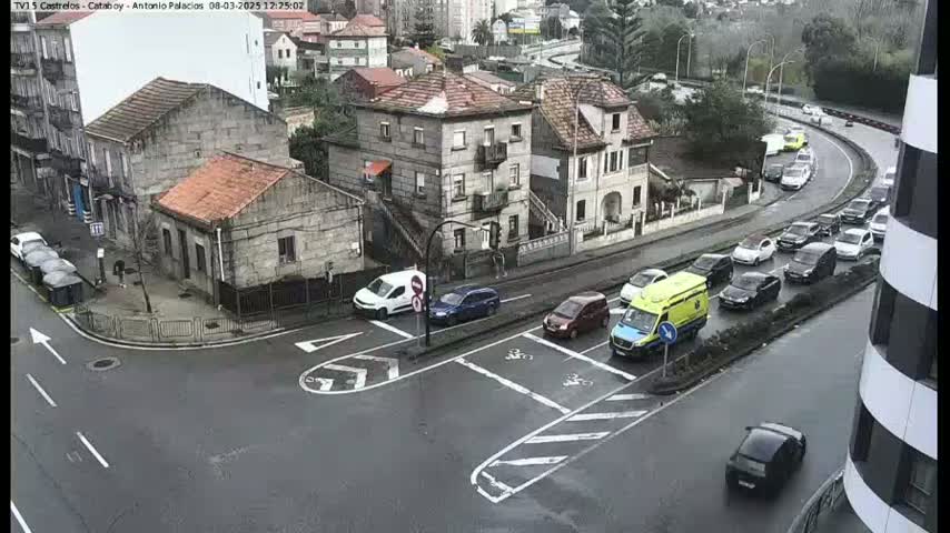A wet, urban intersection with several cars and a van, including an ambulance, at a crossroads near stone buildings.