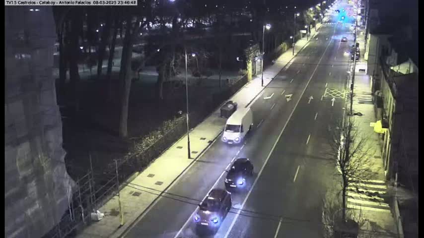 A nighttime, street-level view shows a road with several vehicles including a van and two cars driving down a two-lane road lined with trees and buildings.