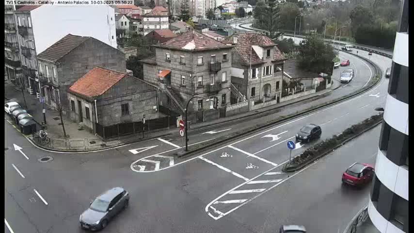 A wet, city intersection with several cars driving on roads around stone buildings.