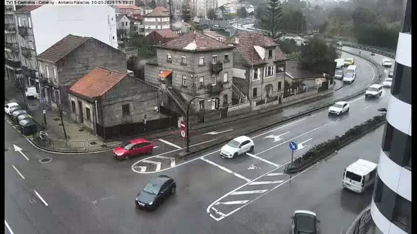 A wet, urban intersection with several cars and buildings, some of which are multi-story.