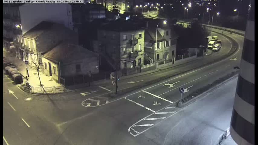 A nighttime street intersection shows several buildings, vehicles, and road markings under artificial light.