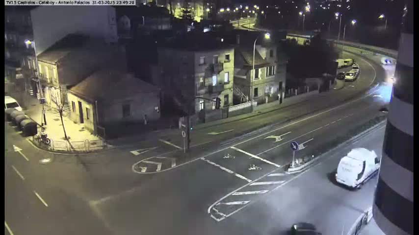 A nighttime aerial view of a street intersection with buildings and vehicles, under mostly clear skies.