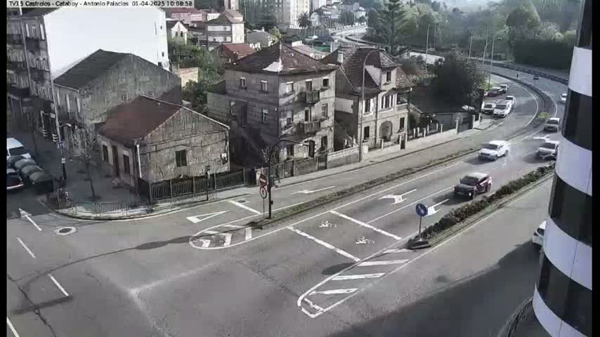 A high-angle, daytime view of a road intersection with several cars driving on a sunny day, near buildings of varying sizes and styles.