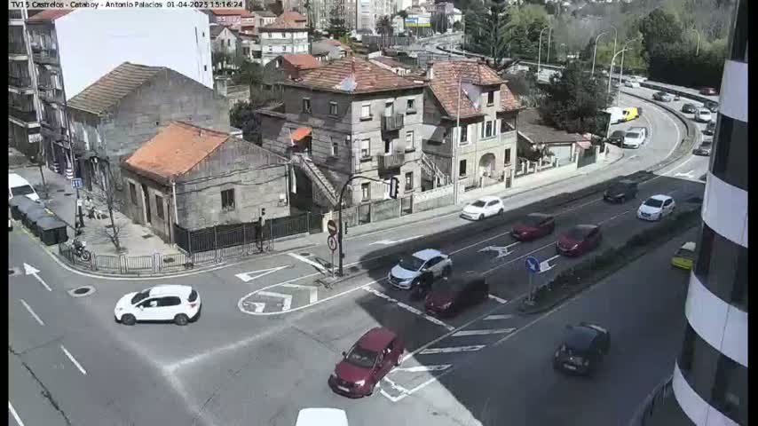 A sunny day shows a street intersection with several cars and buildings, including a multi-story building and several smaller houses.
