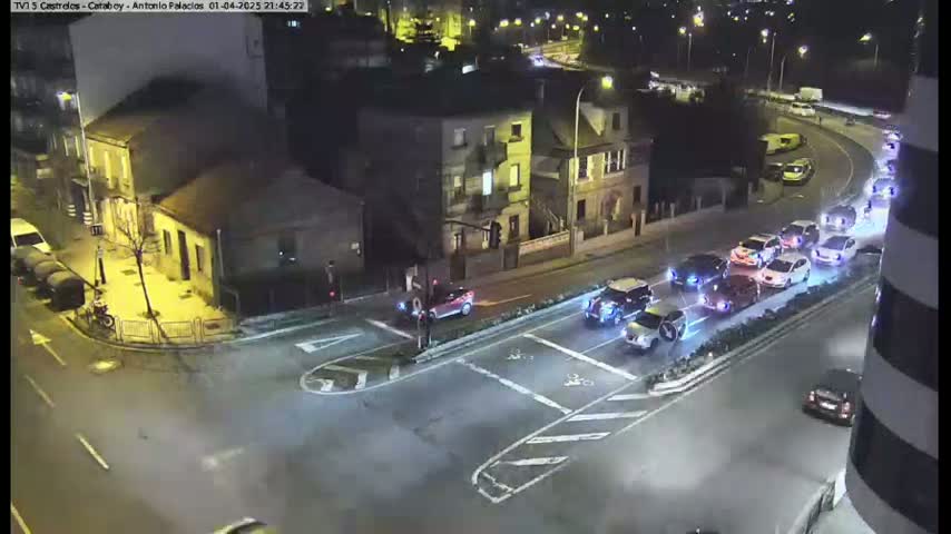 A high-angle nighttime view shows a street intersection with several cars stopped at a traffic light, and more cars driving on a nearby road under artificial street lighting.