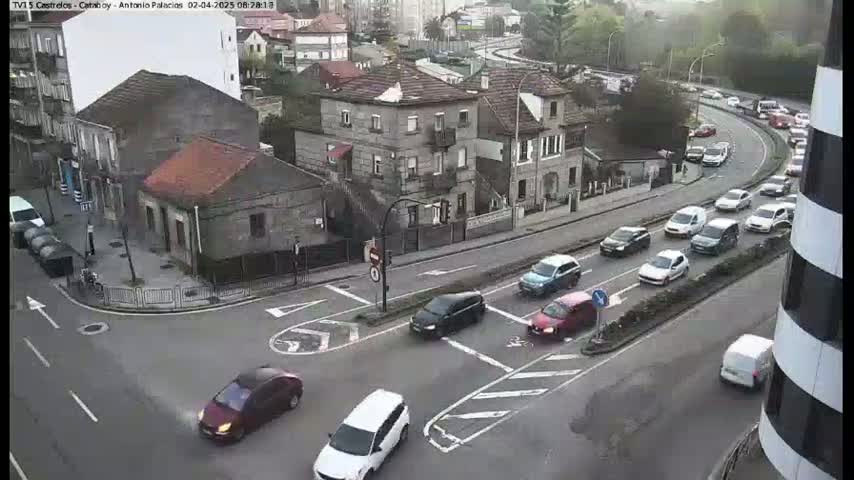 A street intersection with several cars driving on a partly cloudy day, surrounded by buildings.