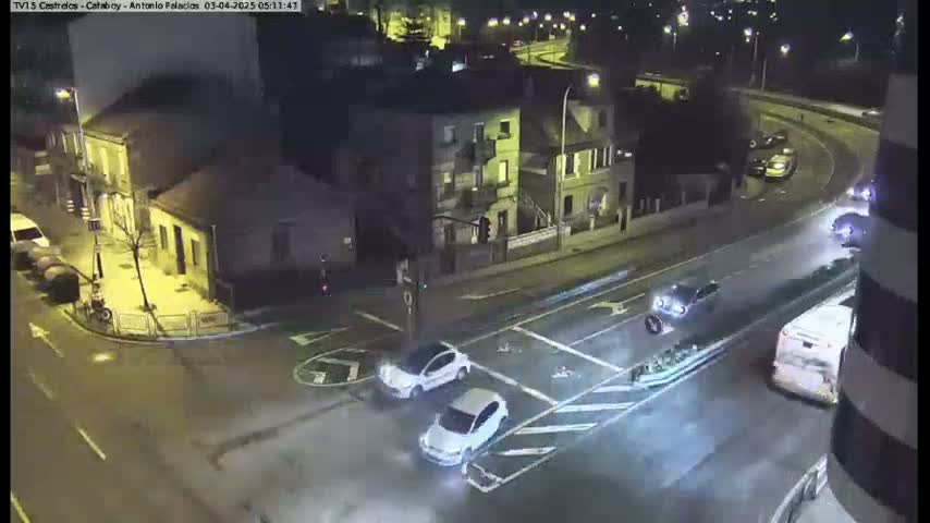 A nighttime street intersection with several cars and a bus, and buildings in the background.