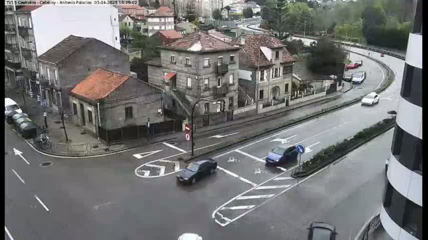 A wet, overcast city intersection shows several cars navigating a multi-lane roadway next to older, multi-story buildings.
