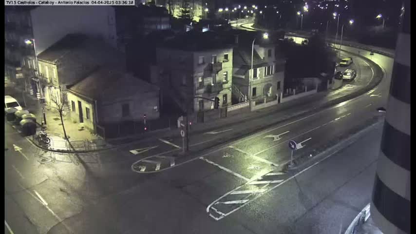 A wet, nighttime street intersection shows several buildings, parked cars, and a few vehicles on the road.