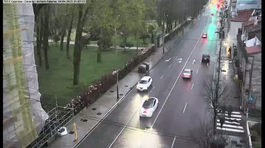 A wet, tree-lined street with several cars driving on it, next to a park and building under construction.
