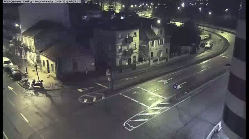 A wet, nighttime street intersection shows several parked cars, a few buildings, and a person on a motorcycle.