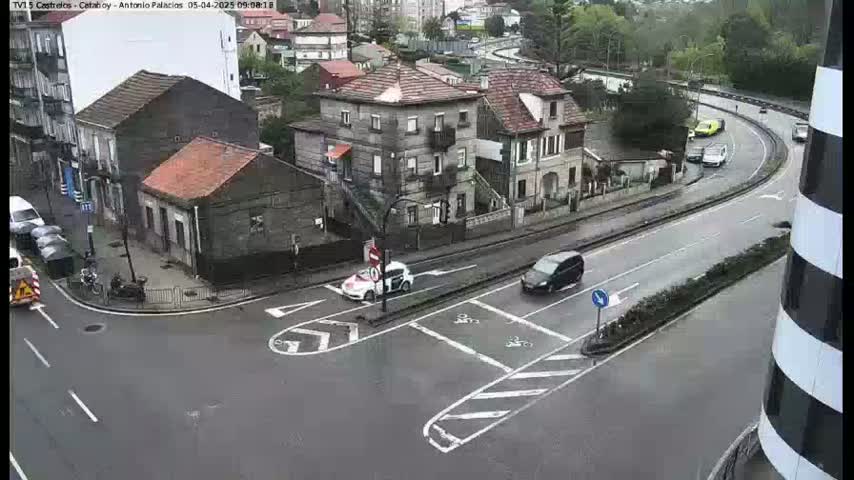 A wet, overcast day shows a street intersection with several cars, buildings, and a curved road.