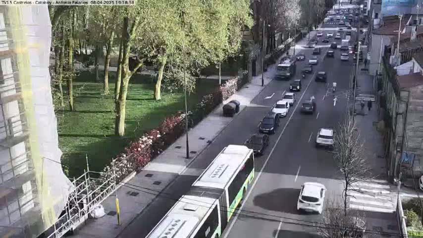 A city street scene on a sunny day shows a long line of cars, a bus, and a partially obscured building under construction, all surrounded by trees and green spaces.