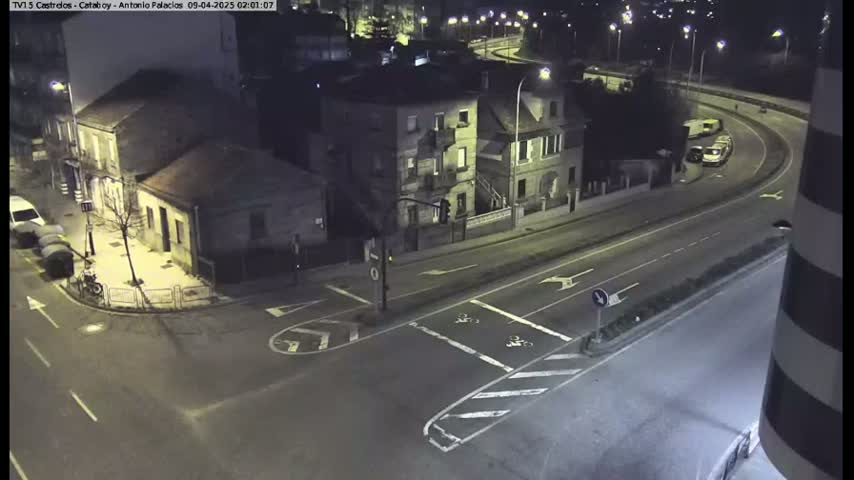 A nighttime, high-angle view shows a street intersection with several buildings and vehicles, including a few parked cars and a couple of trucks on a curved road.