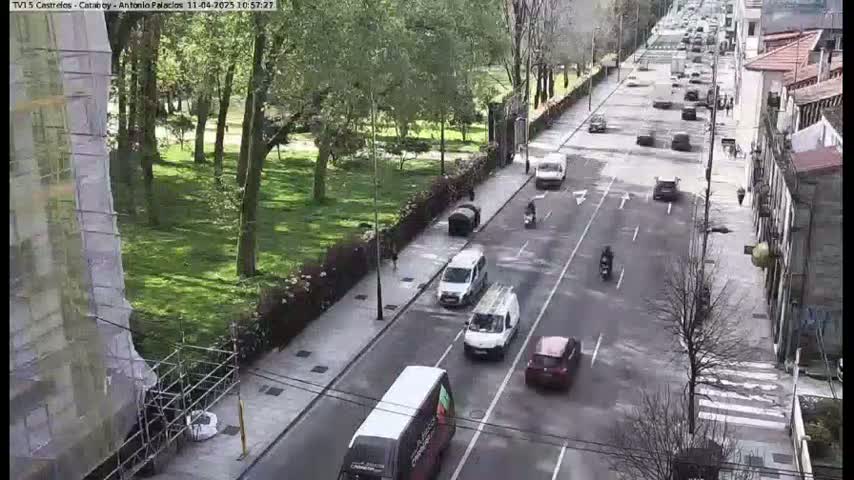 A sunny day shows a city street with several cars and a bus driving along, next to a leafy park with tall trees and buildings under construction.