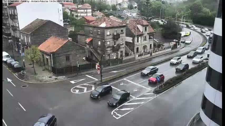 A rainy day shows several cars navigating a street intersection in a city with stone buildings, some parked and others in motion.