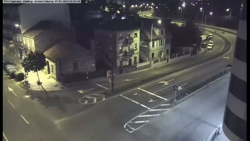 At night, several buildings and a curving roadway are visible, with a few cars and a bicycle present under dimly lit conditions.