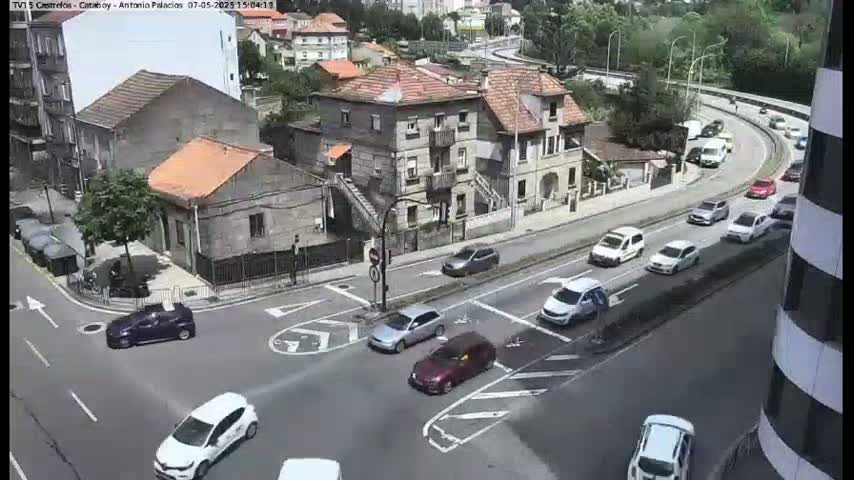 A street intersection with multiple cars, a few buildings, and lush green trees on a sunny day.