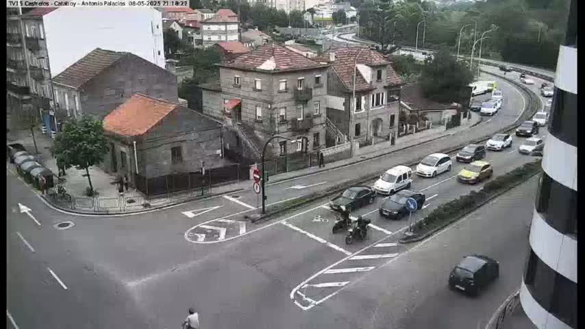 A street intersection in a town shows several cars driving on a roadway that curves to the right, with several buildings and pedestrians visible.