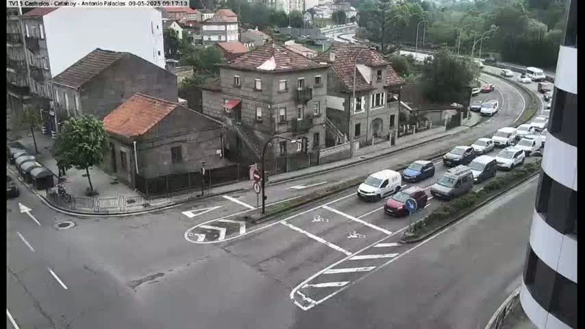 A street intersection in a town with several buildings and many cars, including a line of cars turning onto a curving road, under overcast skies.