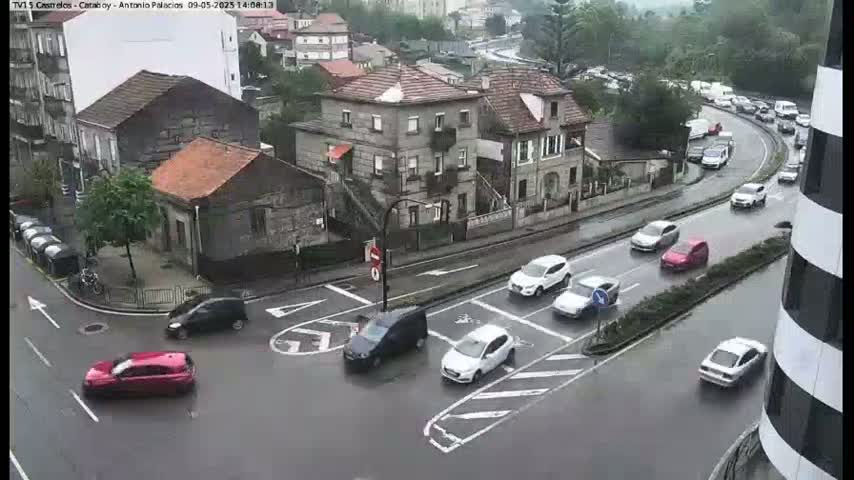 Several cars navigate a wet, city intersection surrounded by buildings under an overcast sky.