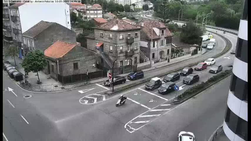 A street intersection with several cars and a motorcycle, showing a partly cloudy day in a town with low buildings.