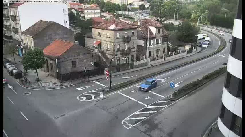 A street intersection with several buildings, a few cars, and a blue car in the foreground on an overcast day.