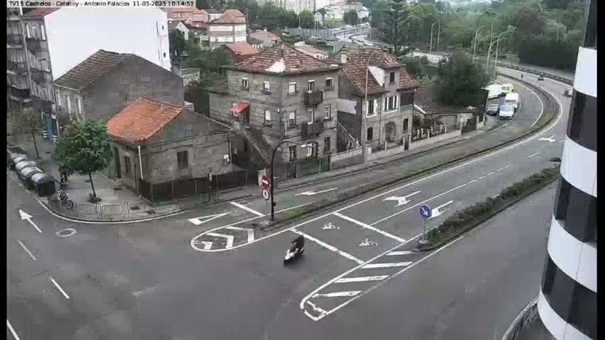 A wet street scene shows a road intersection with several buildings and a few vehicles, including a scooter and some cars on a nearby highway.