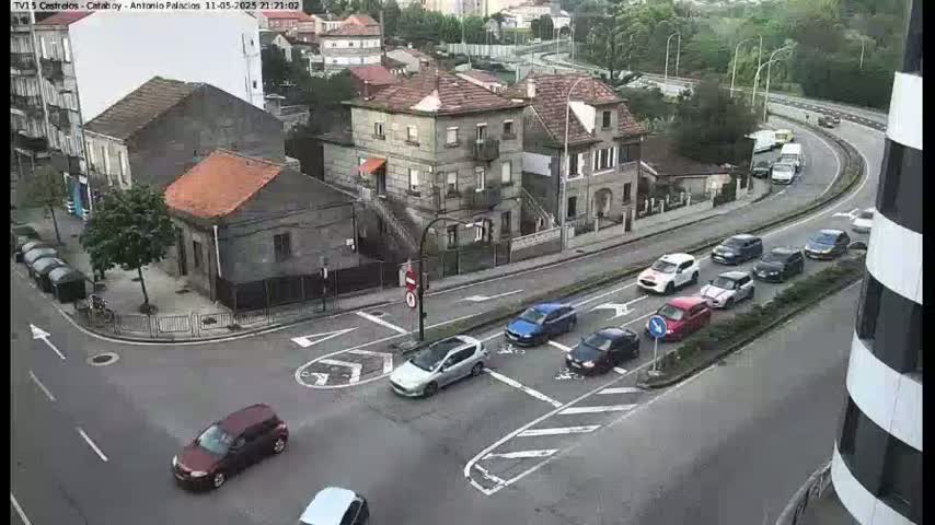 An aerial view shows a street intersection with several cars at various points, and several buildings including multi-story structures, on an overcast day.