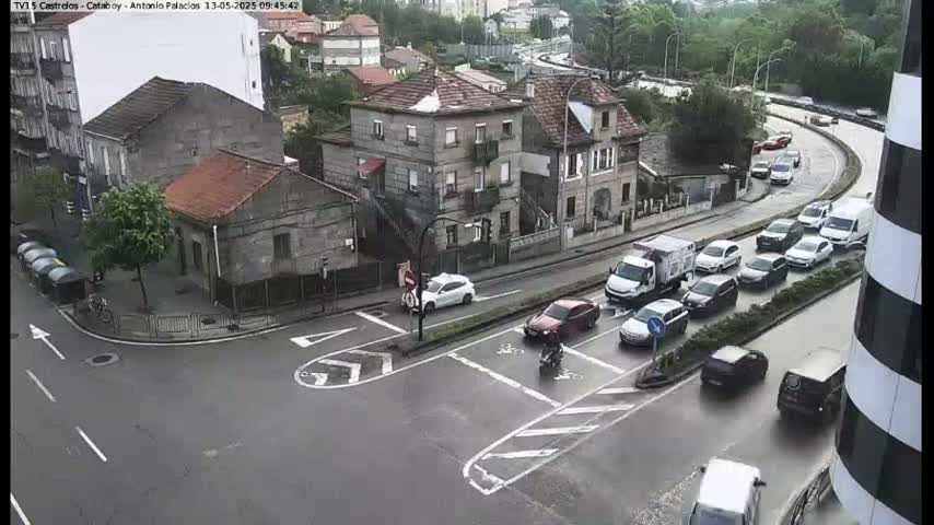 A wet, overcast day reveals a street intersection with several cars stopped at traffic signals and several more cars driving on a nearby, curving roadway, all surrounded by stone buildings.