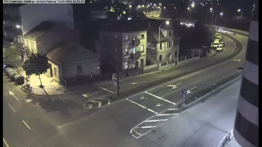 At night, a mostly empty road curves through a residential area with buildings and streetlights illuminating the scene.