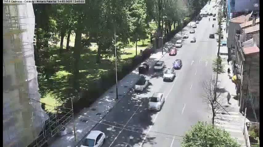 A sunny day shows a city street with several cars driving in both directions, lined with trees and buildings, one of which is under construction.