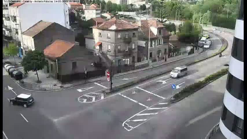 A street intersection with several buildings and cars, including a small dark car turning left, a white van moving forward, and other vehicles parked nearby, under a partly sunny sky.